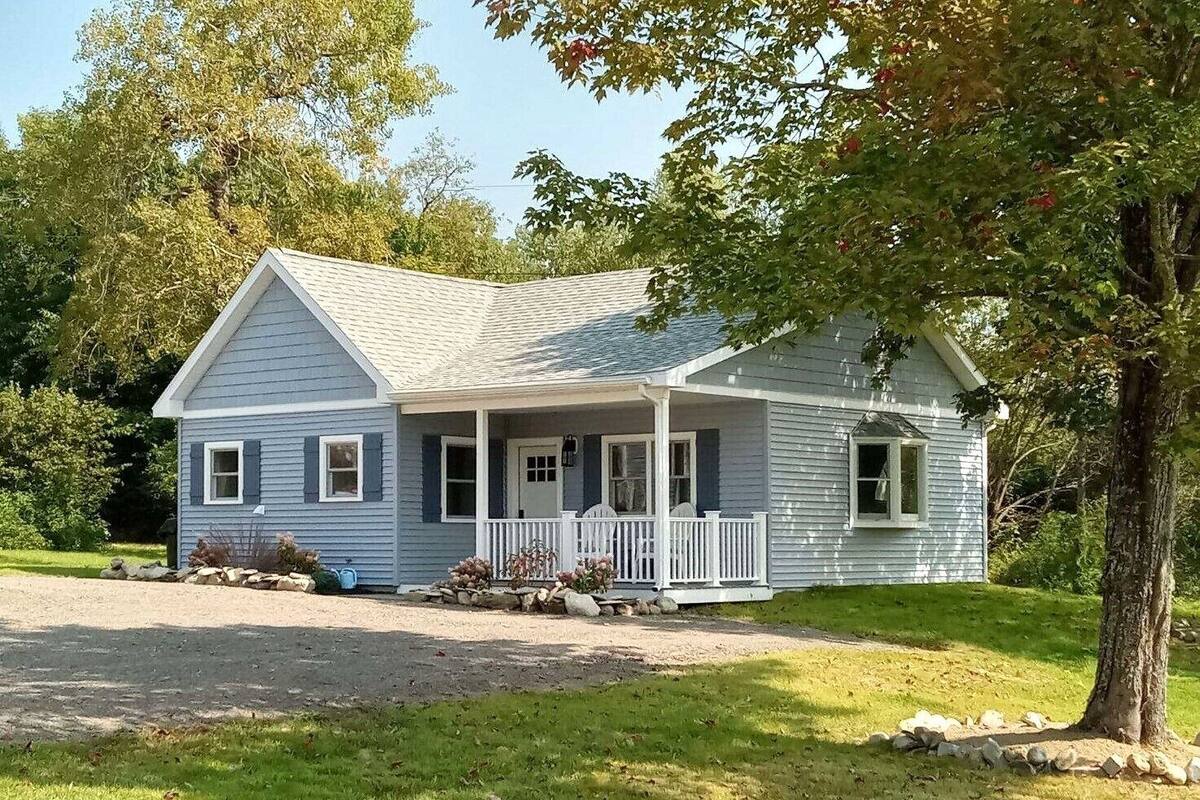 A single-story house with a gabled roof, a front porch with white railings, and a small front yard with a tree and a stone wall.