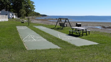A grassy area with a hopscotch court, a swing set, and a picnic table by the beach.