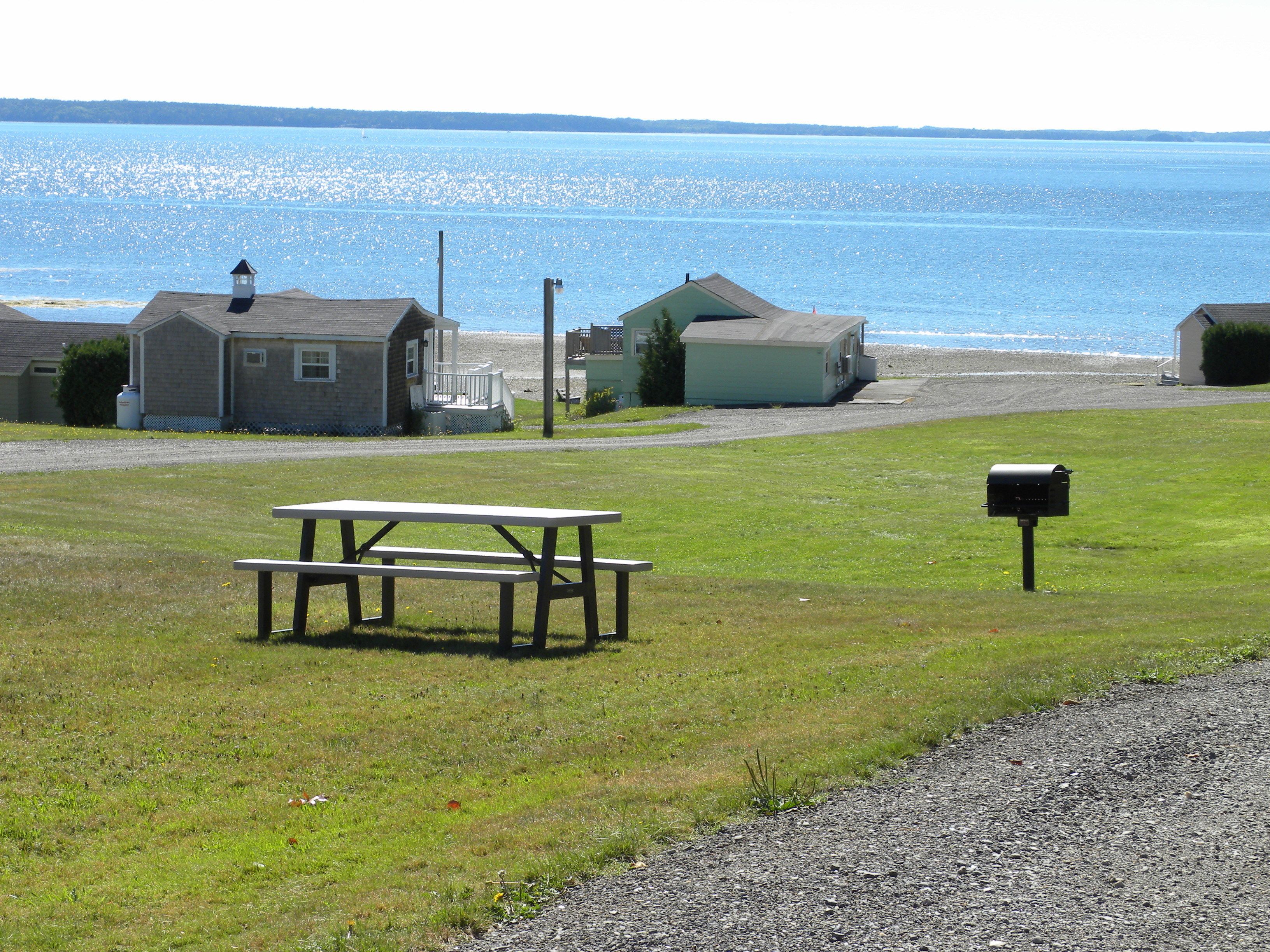 A picnic table and a mailbox in a grassy area with houses and a beach in the background.