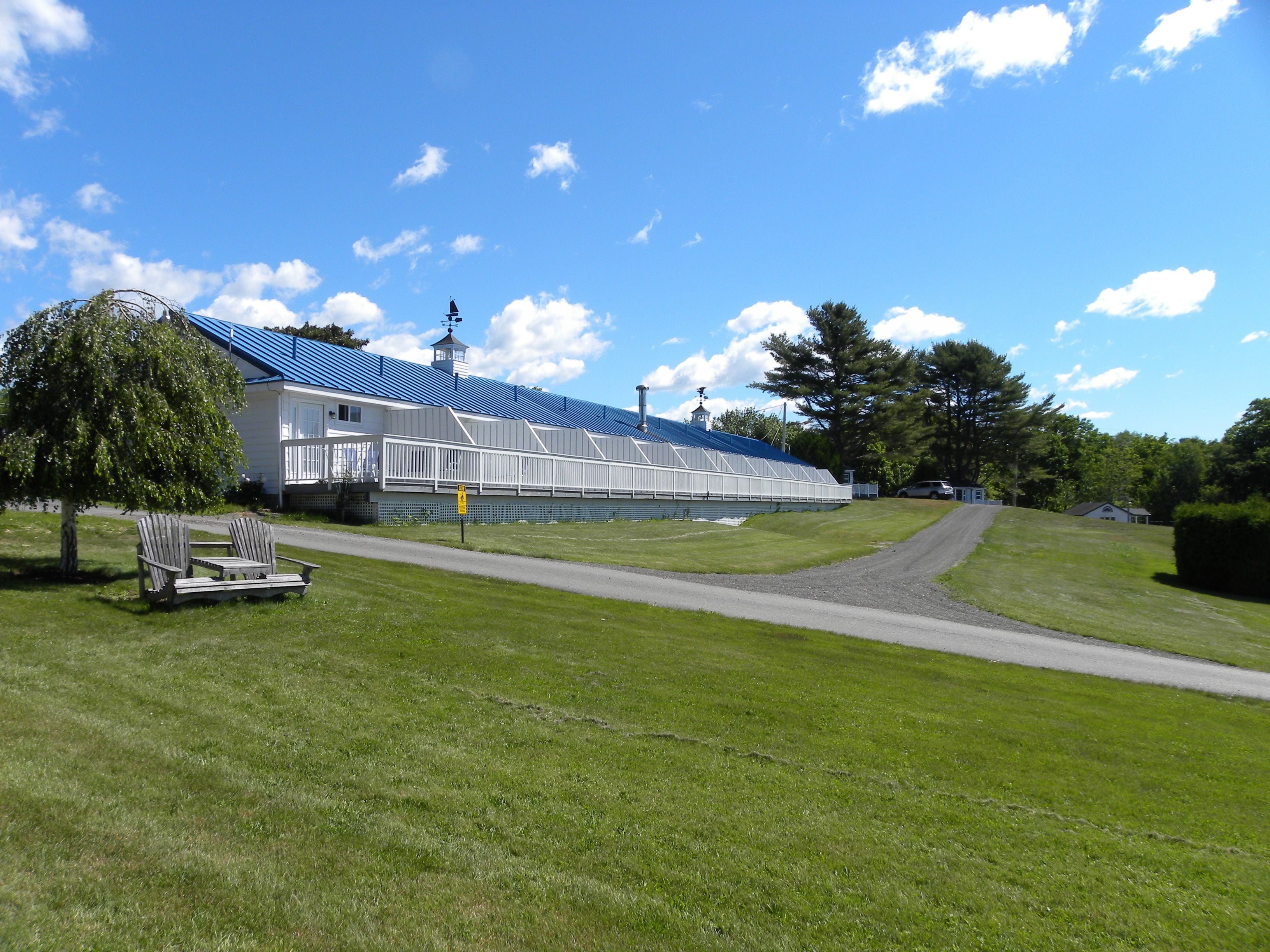 A large, white building with a blue roof, a covered walkway, and a grassy area with two Adirondack chairs.