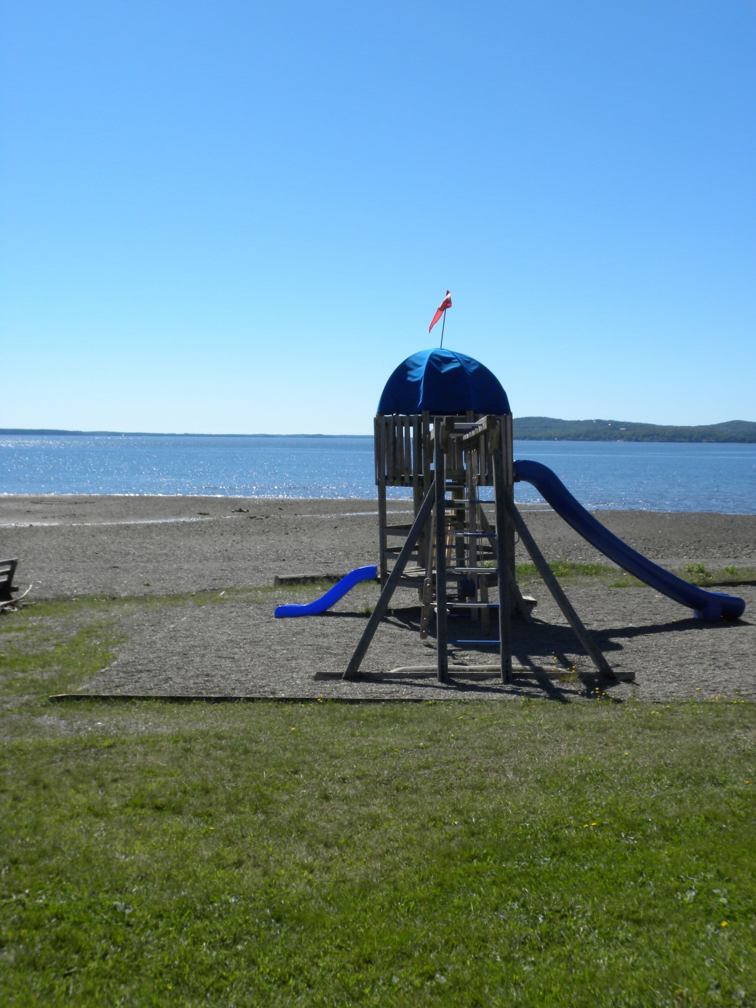 Ein Spielplatz mit einer Rutsche und einer blauen Markise, der sich auf einer Wiese in der Nähe von Gewässer befindet.