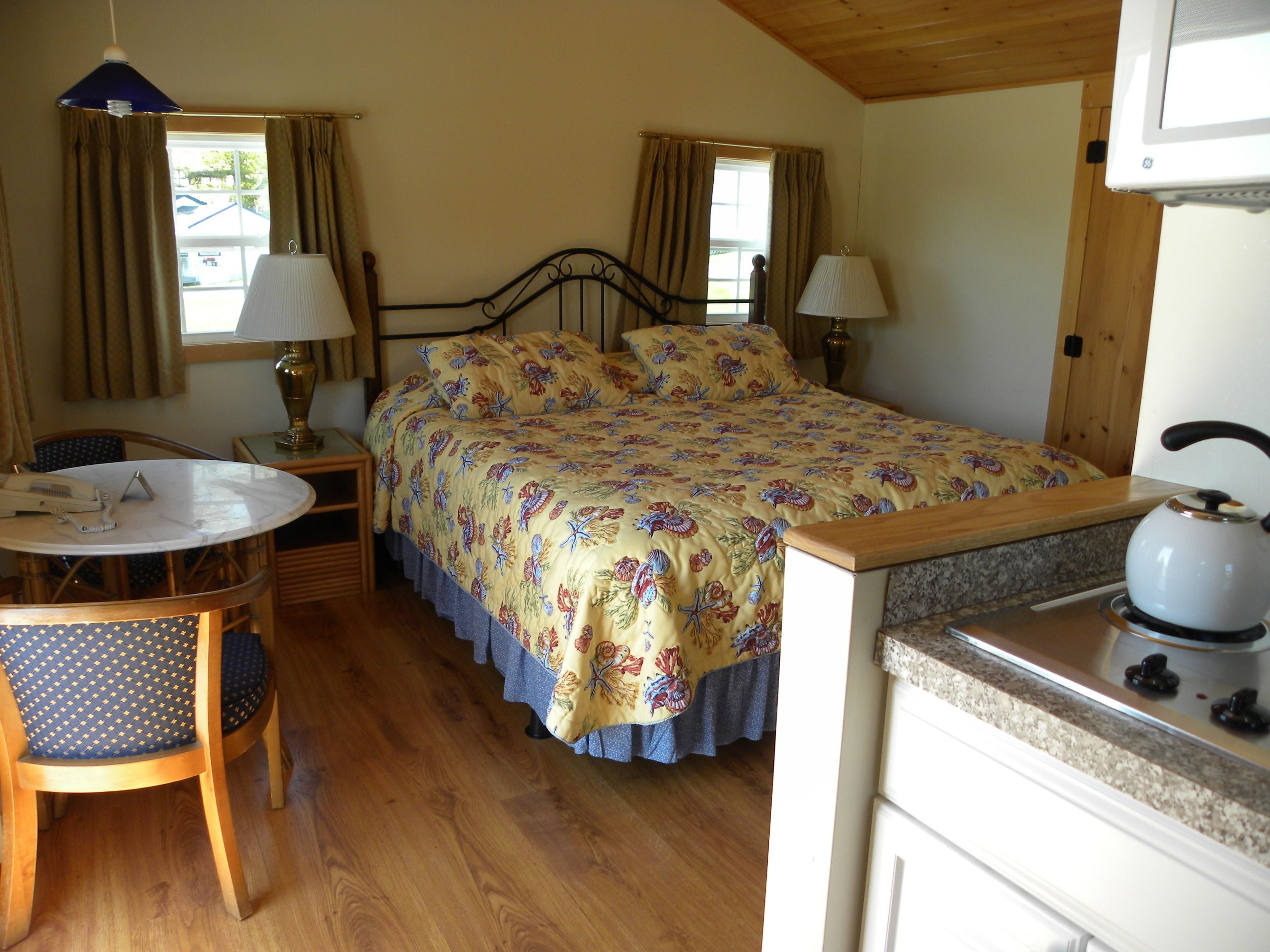 A cozy bedroom with a bed, bedside lamps, a small table with a chair, and a kitchenette.