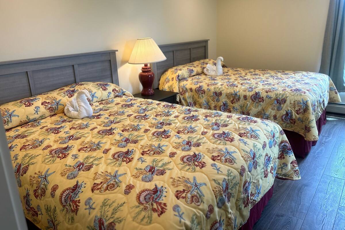 Two beds with floral and sea creature bedding, a nightstand with a lamp, and a window with curtains.