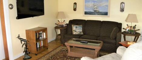 A living room with a brown sofa, a white armchair, a coffee table, a television mounted on the wall, a bookshelf, and a painting of a ship on the wall.