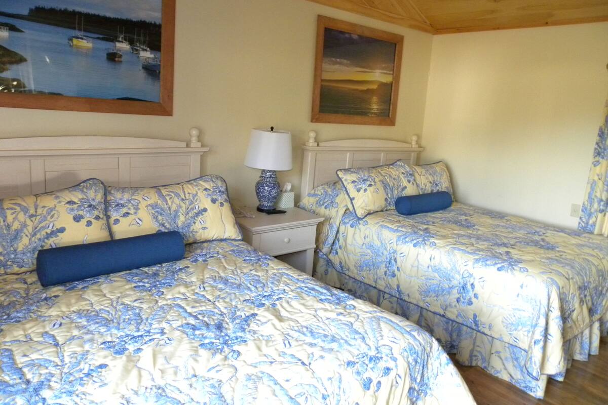 A room with two beds, each with floral bedding and blue pillows, a nightstand with a lamp, and a painting of a harbor scene on the wall.