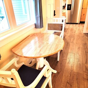 A dining area with a wooden table and white chairs, a window with blinds, and a kitchen in the background.