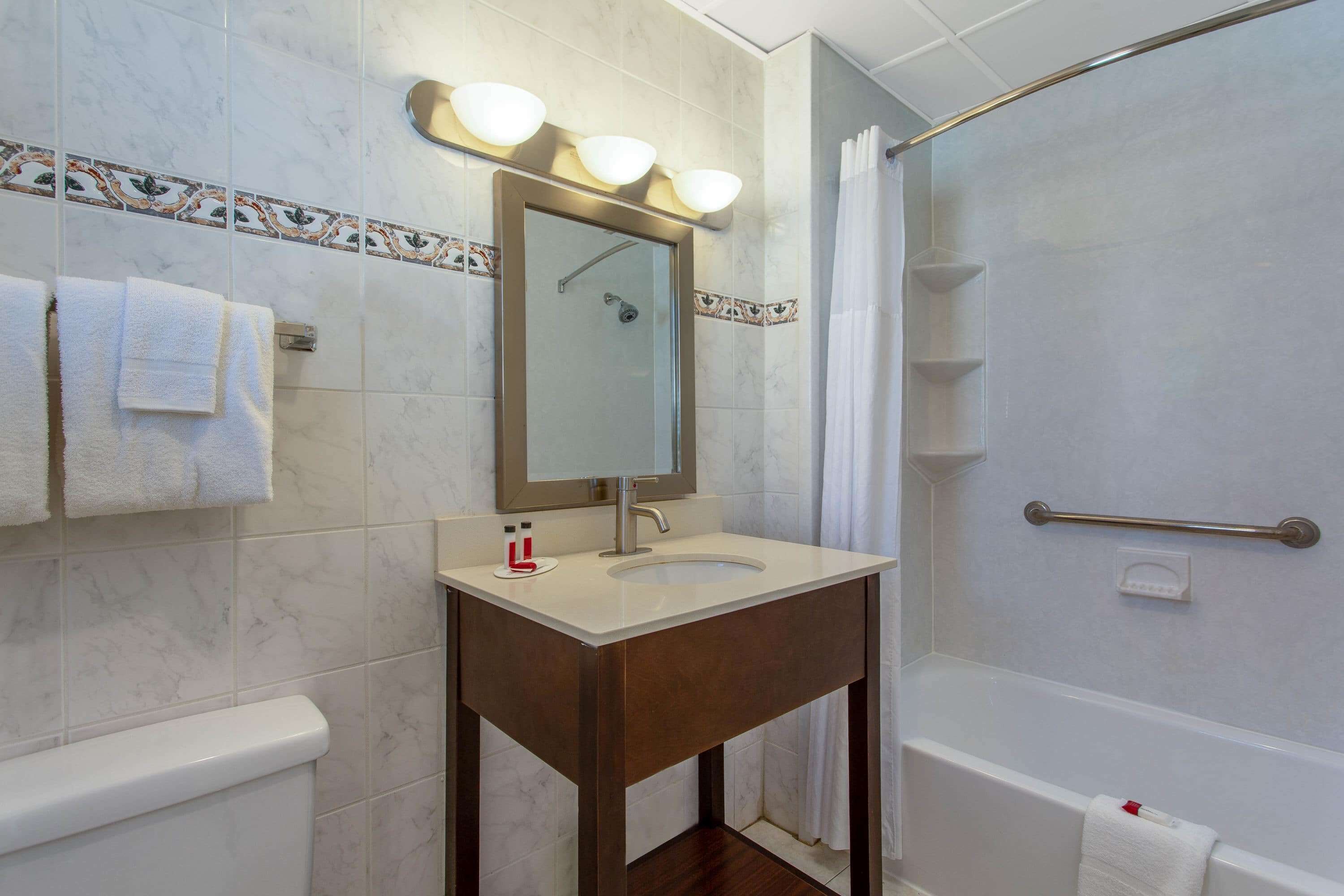 A bathroom with a white sink, mirror, and shower. There are towels on the wall and a handrail in the shower.