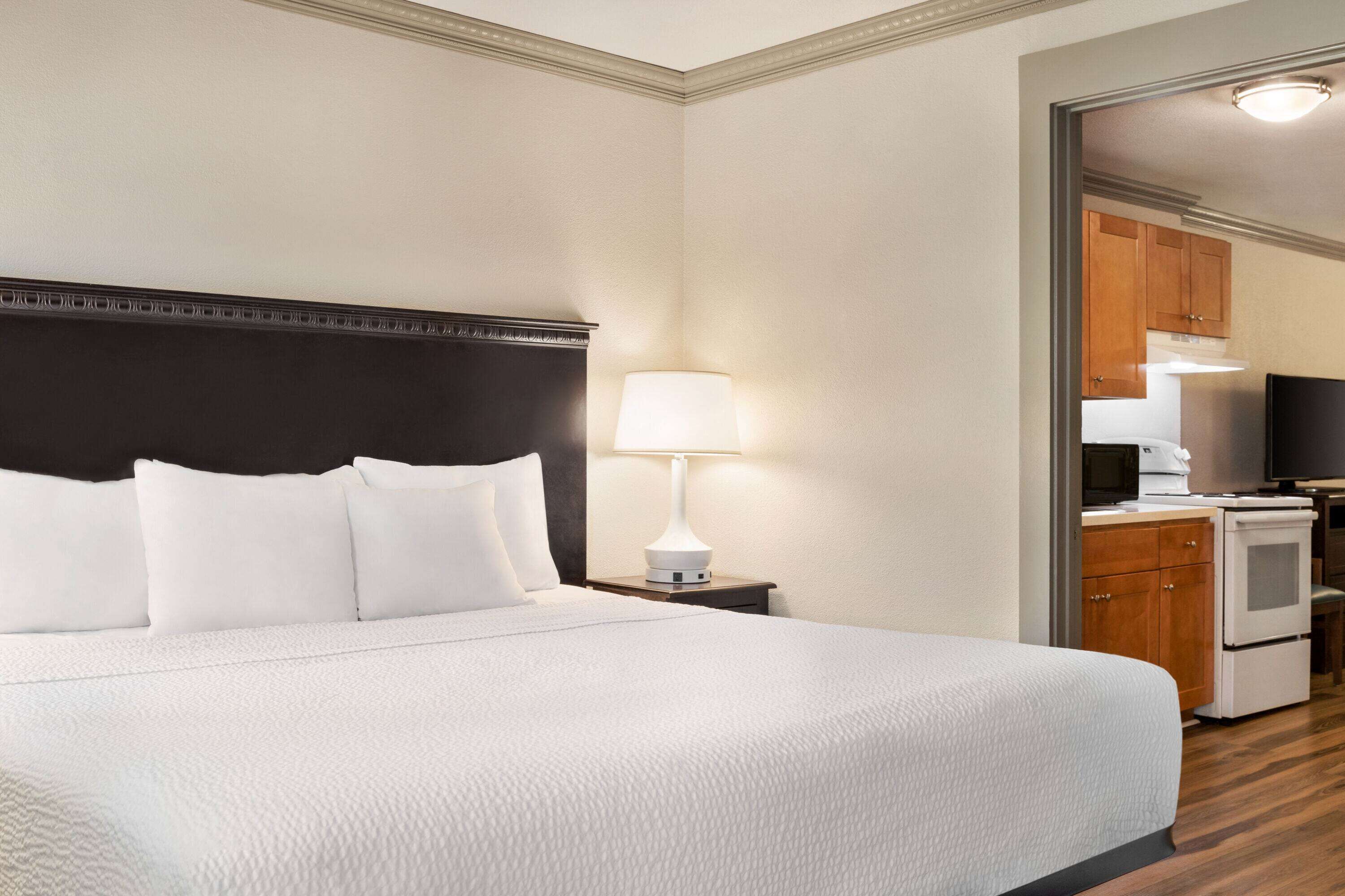 A hotel room with a large bed, a bedside lamp, a small table, and a kitchenette in the background.