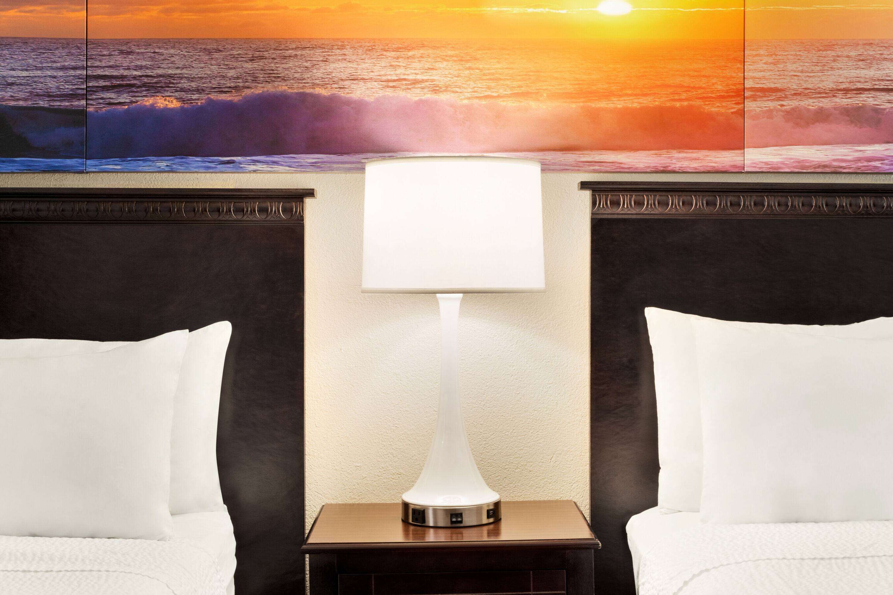 Habitación de hotel con cama, mesita de noche con lámpara y un mural en la pared que muestra un atardecer sobre el océano.