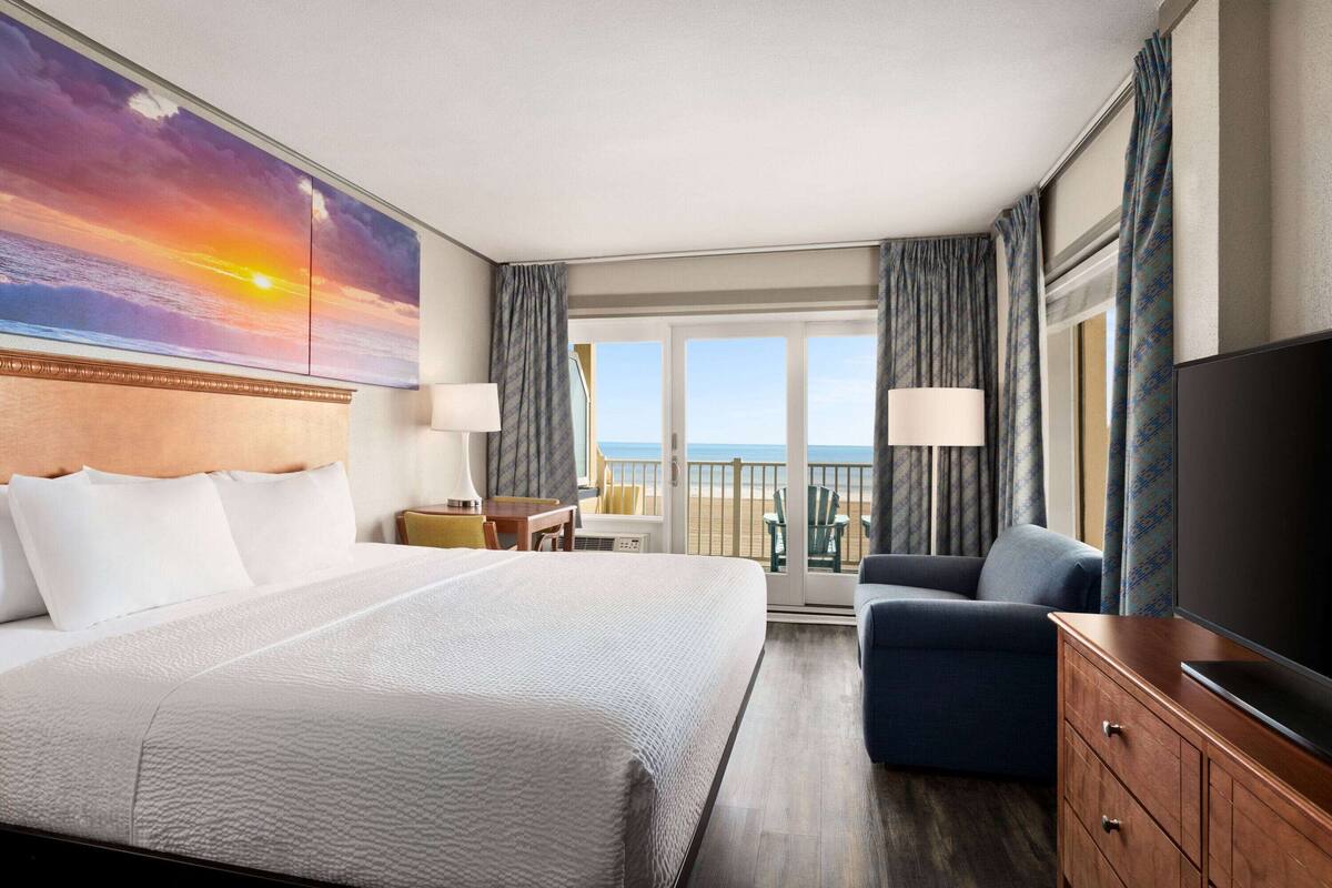 A hotel room with a large bed, a TV, a desk, and a balcony with a view of the ocean.