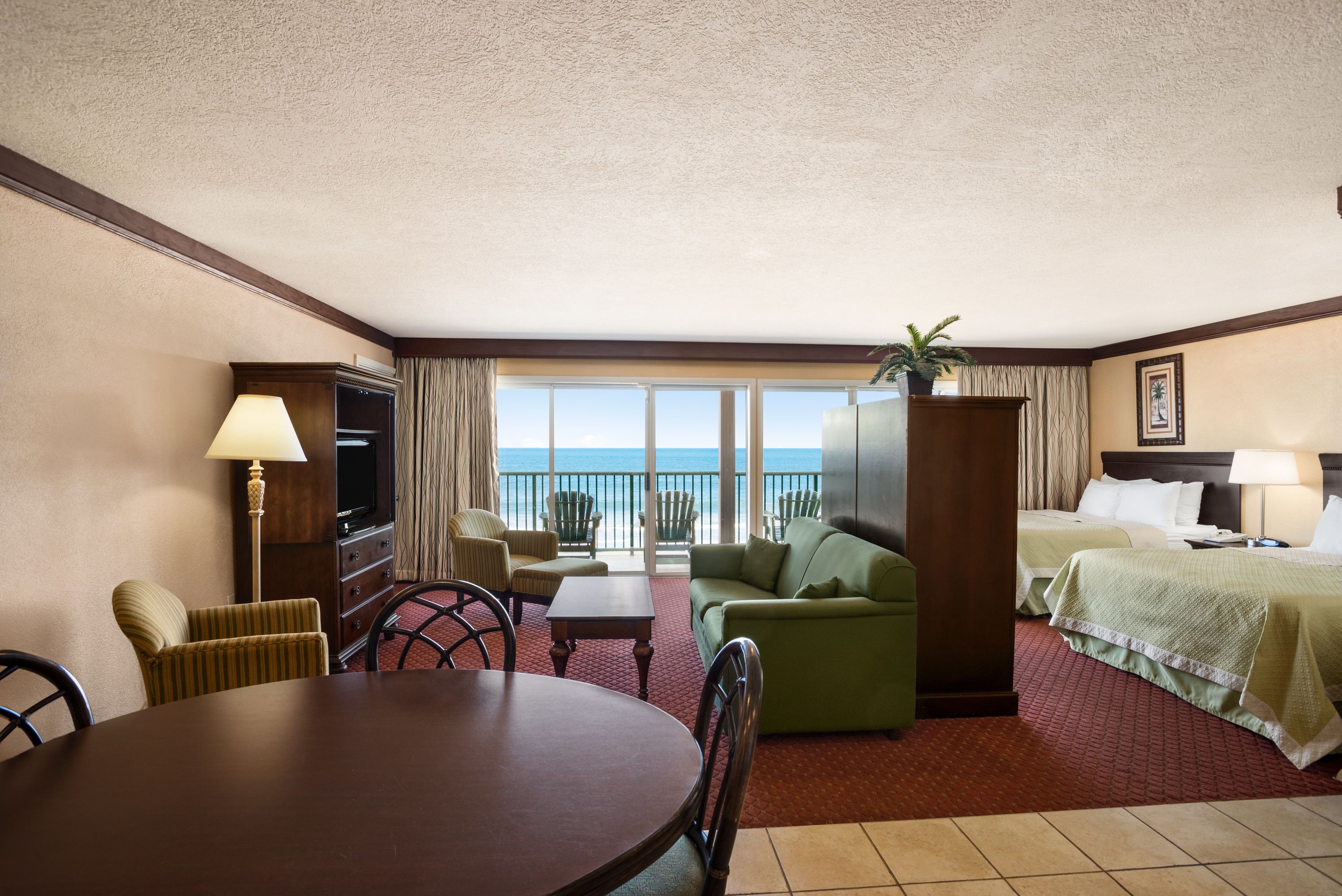 A hotel room with a dining table, green sofa, two beds, a TV, and a view of the ocean.