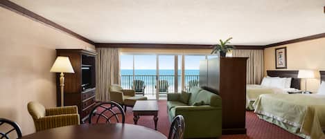 A hotel room with a dining table, green sofa, two beds, a TV, and a view of the ocean.