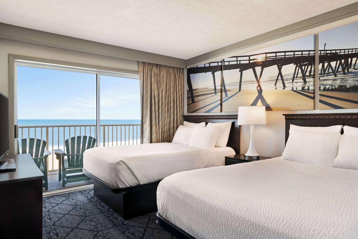 A hotel room with two beds, a desk, a chair, and a large window with a view of a pier and beach.