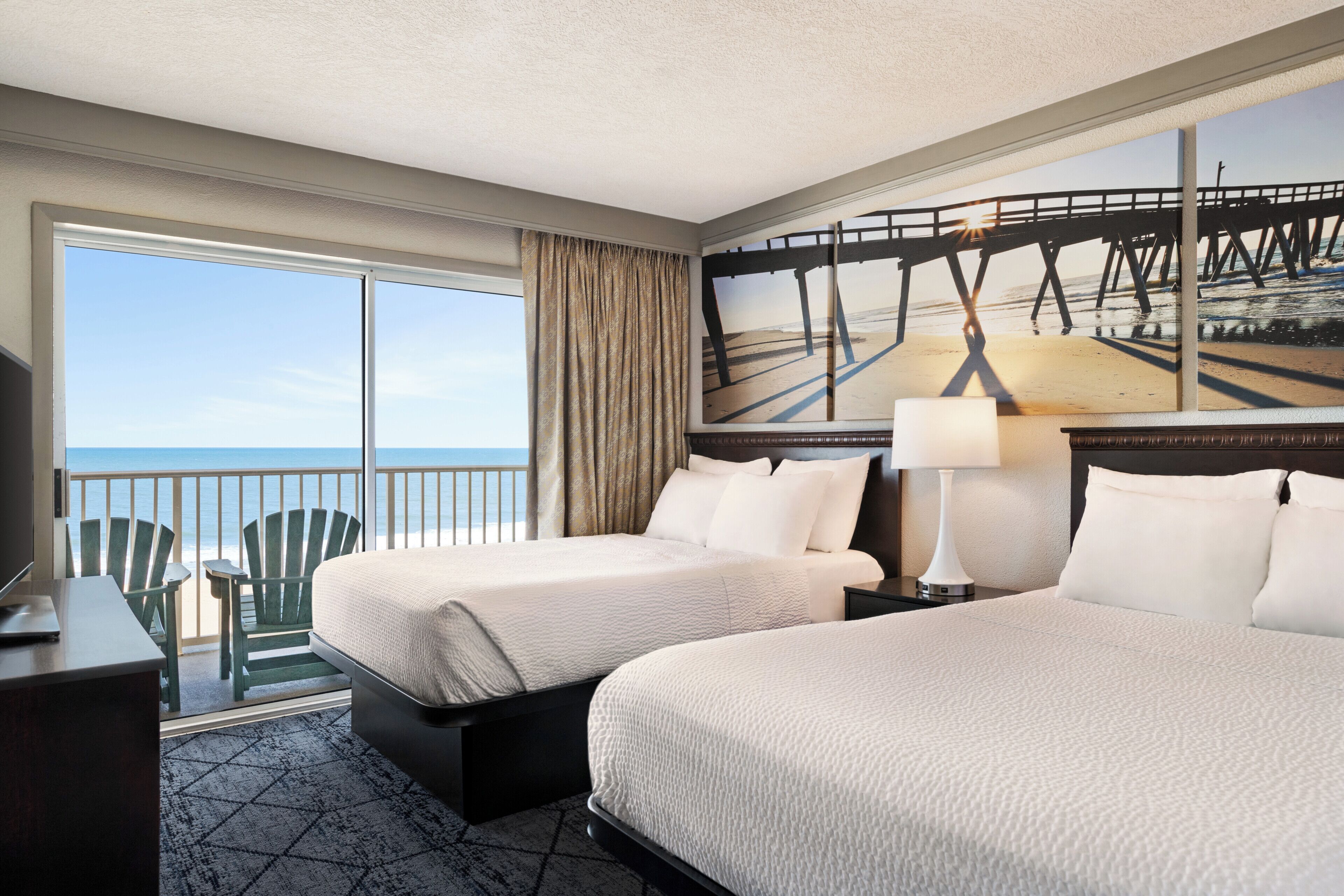 Ein Hotelzimmer mit zwei Betten, einem Schreibtisch, einem Stuhl, einem großen Fenster mit Meerblick und einem Gemälde eines Piers.