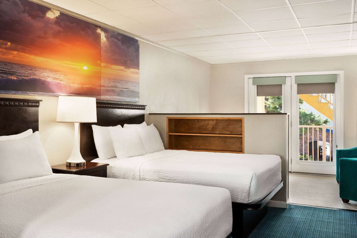 A hotel room with two beds, a nightstand with a lamp, a large painting of a sunset, a wooden headboard, and a balcony with a view.