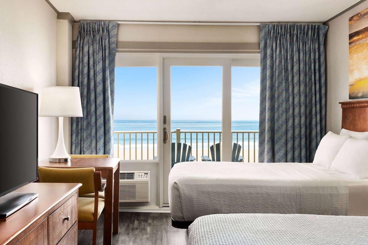 A hotel room with a bed, a desk with a lamp, a TV, and a view of the beach.