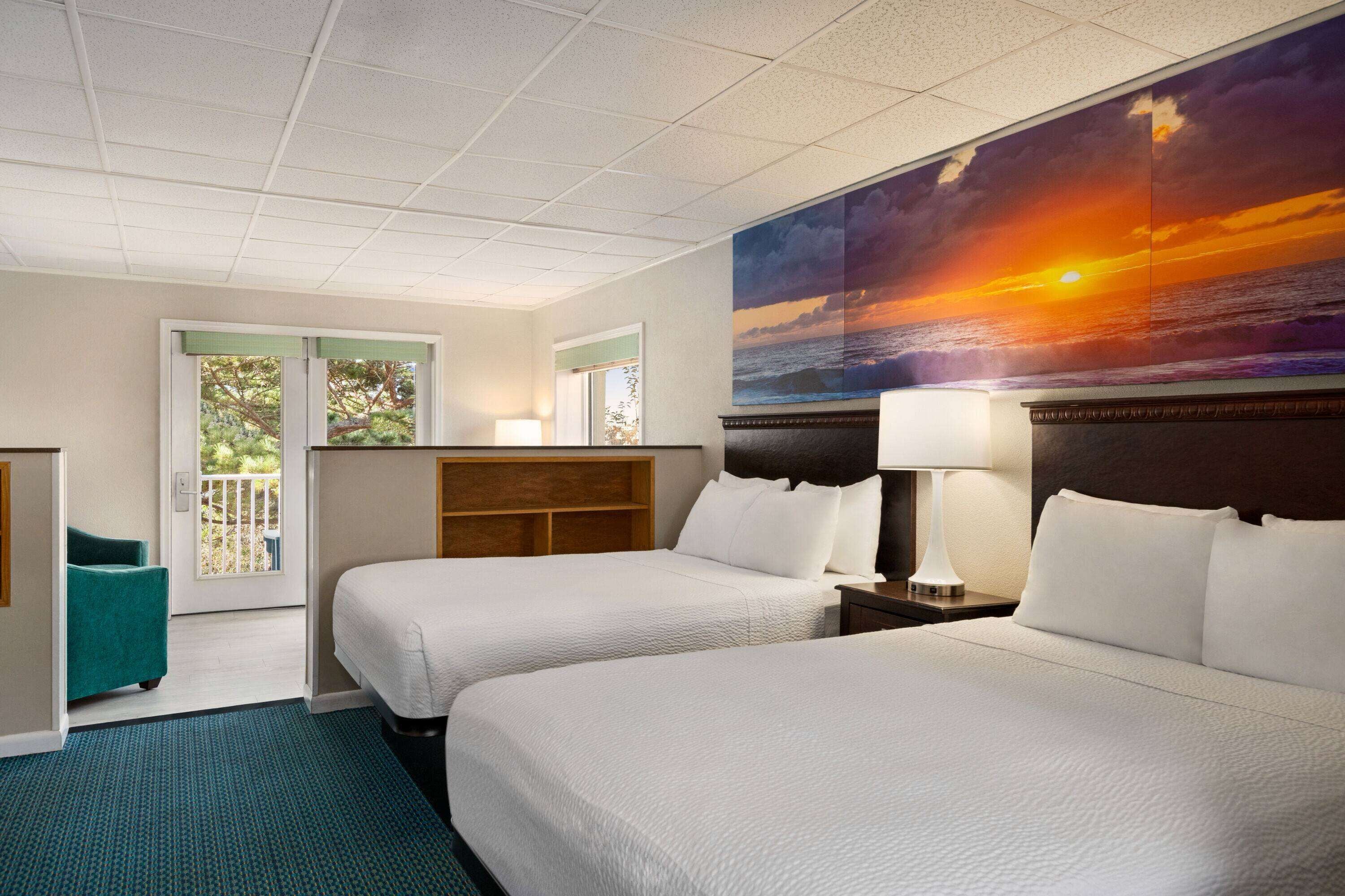 A hotel room with two beds, a large painting of a sunset, a view of the outdoors, and a teal chair.