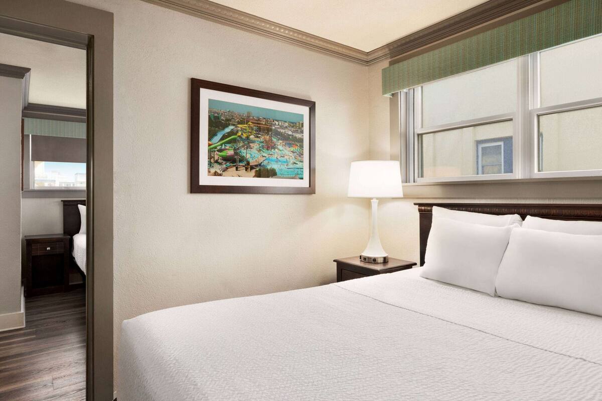A hotel room with a large bed, a framed picture on the wall, a bedside lamp, and a window with blinds.