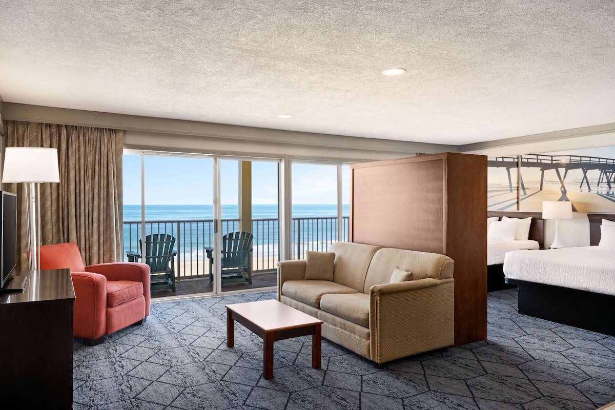 A hotel room with a balcony, a sofa, a chair, a bed, and a desk with a lamp.
