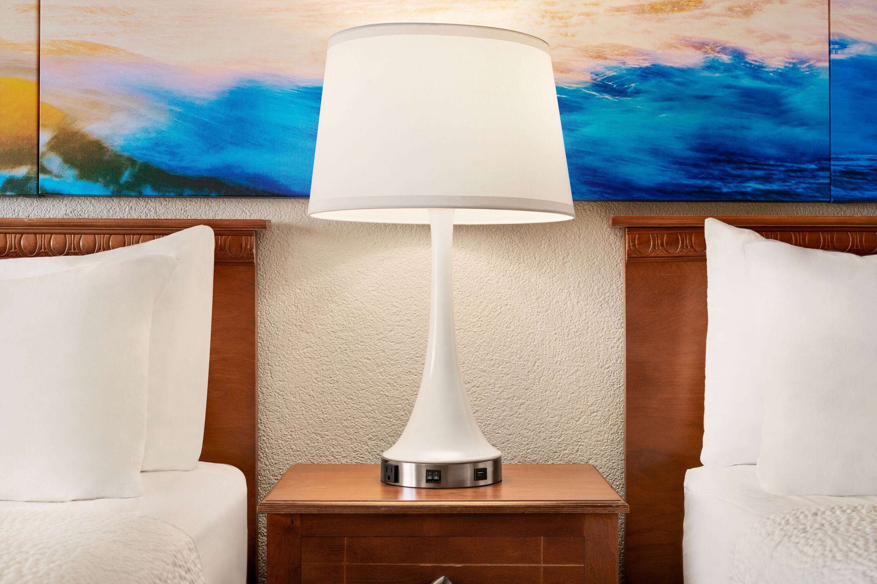 Una camera d'albergo moderna con una lampada bianca su un comodino in legno, un quadro grande appeso al muro e due cuscini bianchi su una testiera in legno.