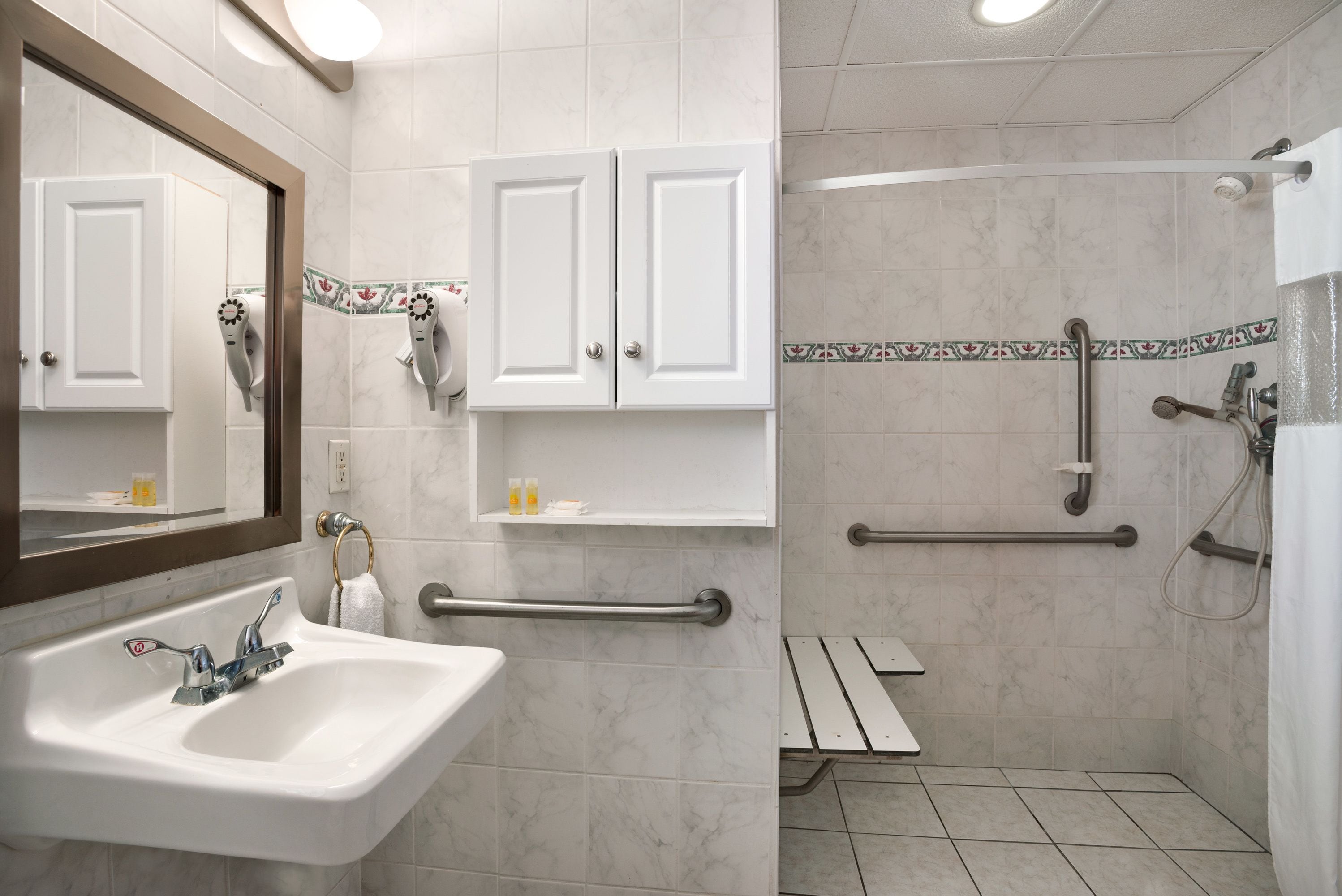 A bathroom with a sink, two cabinets, a mirror, a shower area with grab bars, and a towel rack.