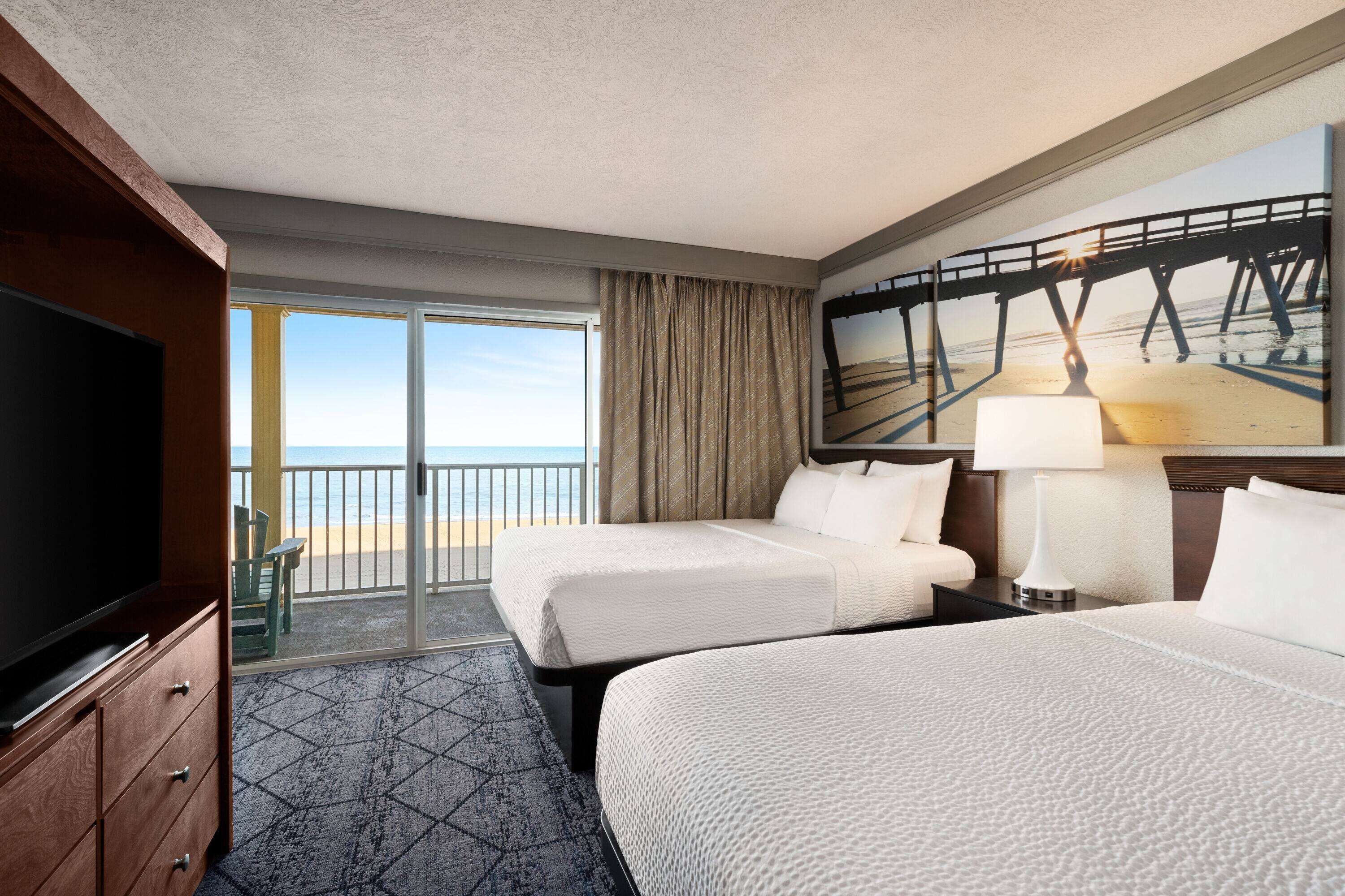 A hotel room with two beds, a TV, a painting of a pier, and a balcony with a view of the beach.