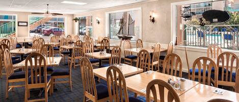 A restaurant with multiple wooden tables and chairs, large windows with a view of a swimming pool and buildings, and ceiling fans.
