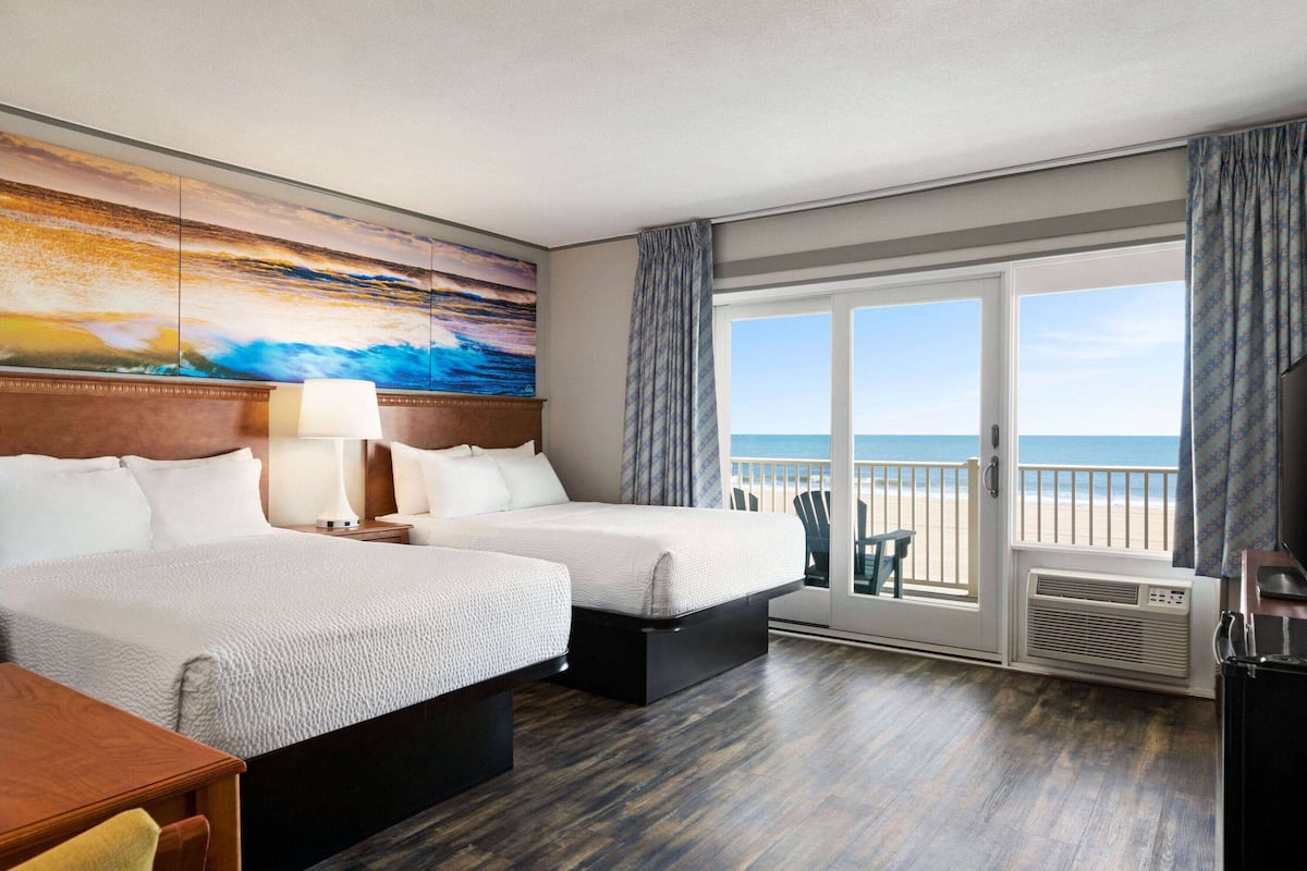A hotel room with two beds, a desk, a chair, a painting, a view of the beach, and a sliding door to the balcony.