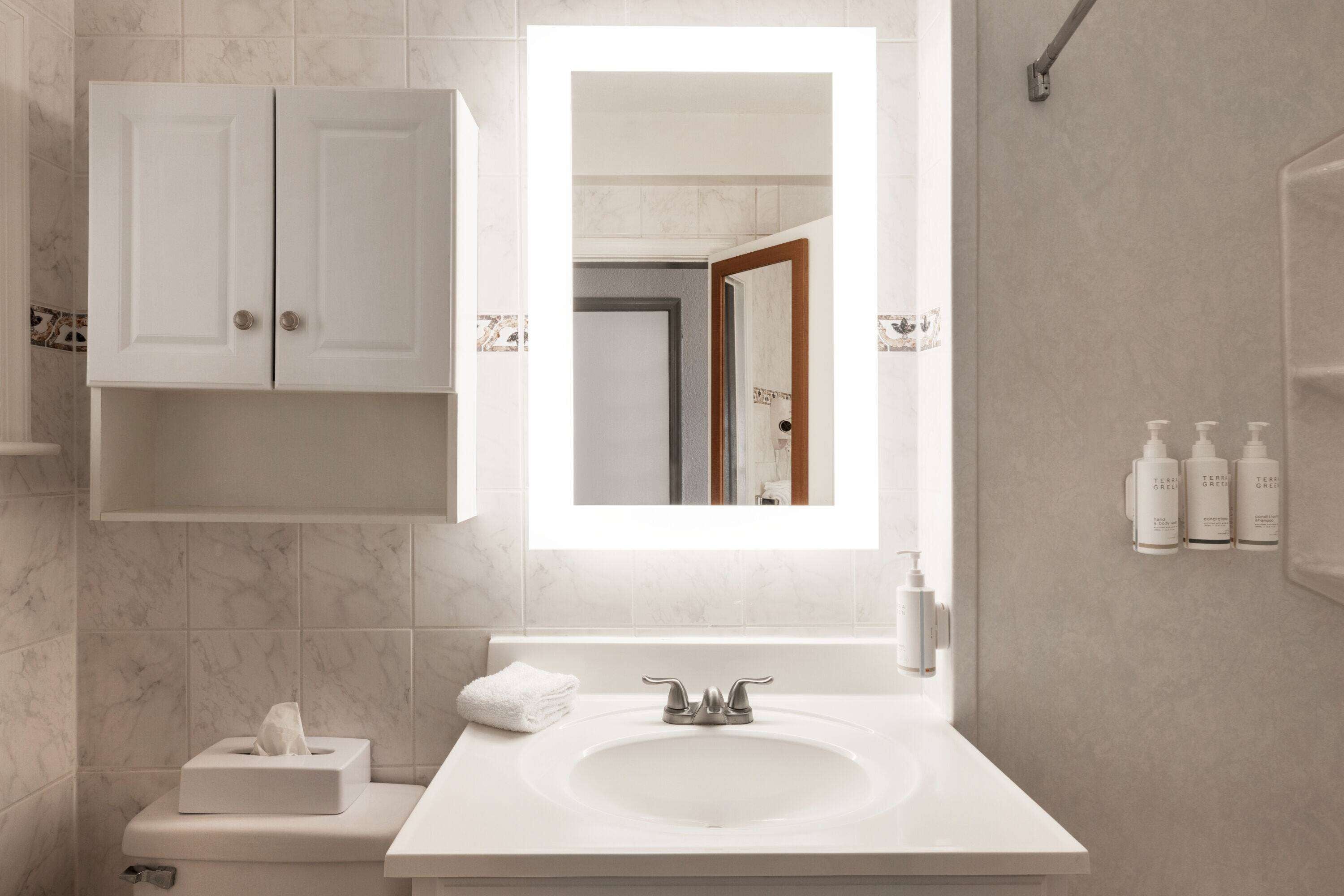 A modern bathroom with a white sink, a mirror above it, and a toilet with a tissue box. There are toiletries on the wall and a towel on the floor.