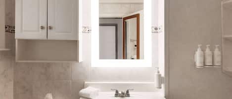 A modern bathroom with a white sink, a mirror above it, and a toilet with a tissue box. There are toiletries on the wall and a towel on the floor.