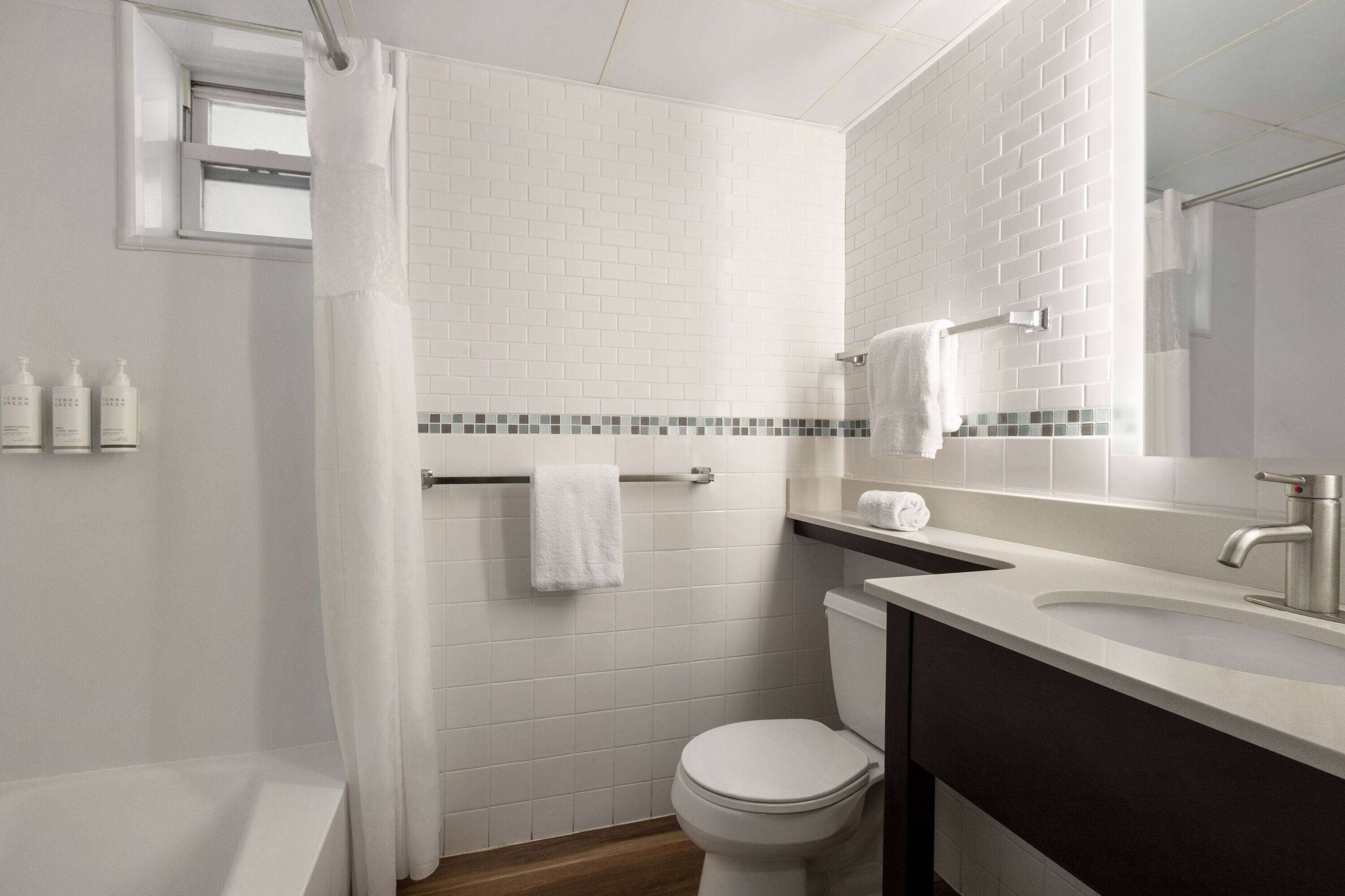 Et moderne badeværelse med en hvid flisevæg, trægulv, toilet, badekar, vaskebord med spejl og et håndklædetørrestativ.