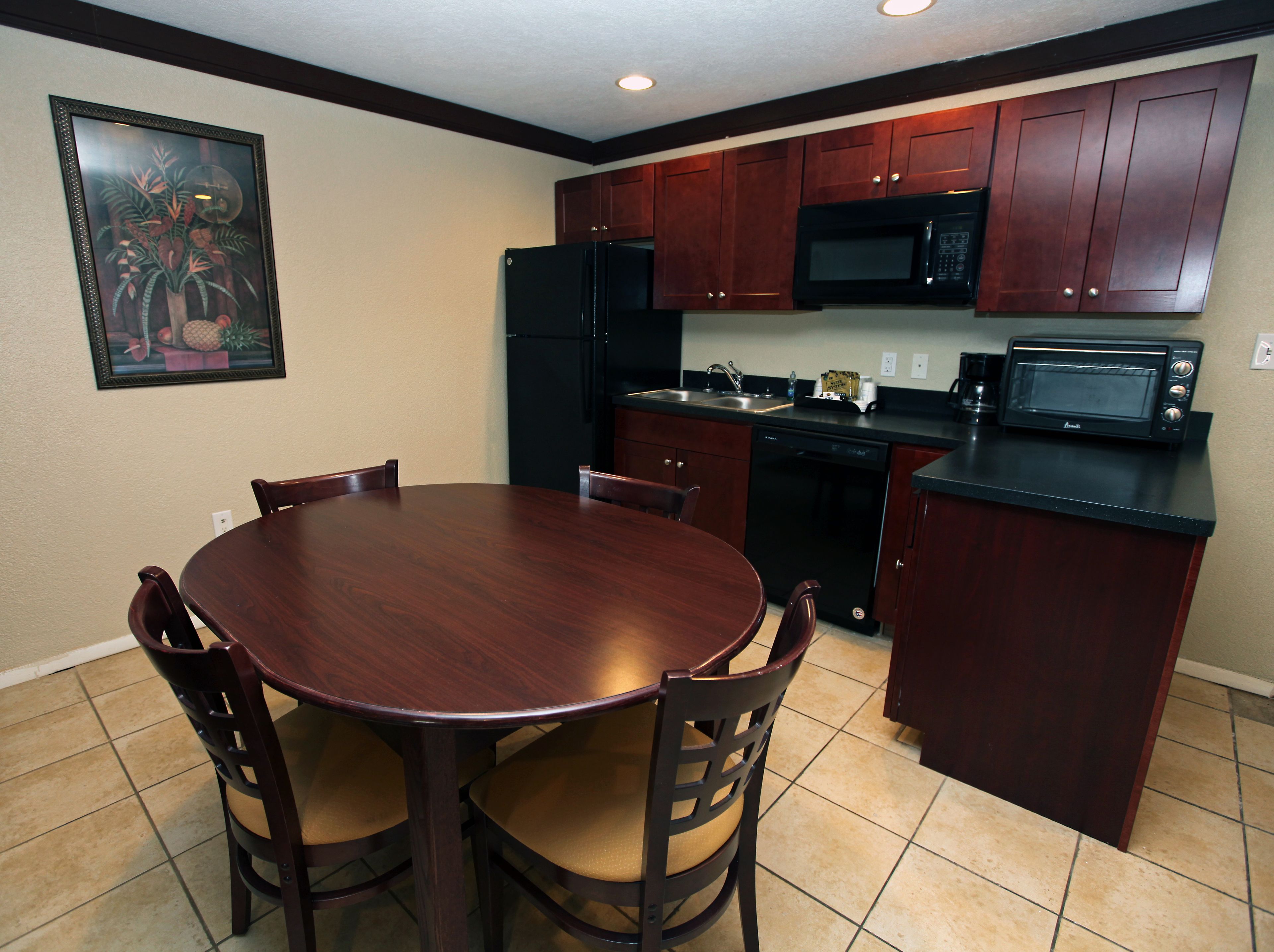 A kitchen with dark wooden cabinets, a black refrigerator, a microwave, and a dishwasher. There is a dining table with chairs and a framed picture on the wall.