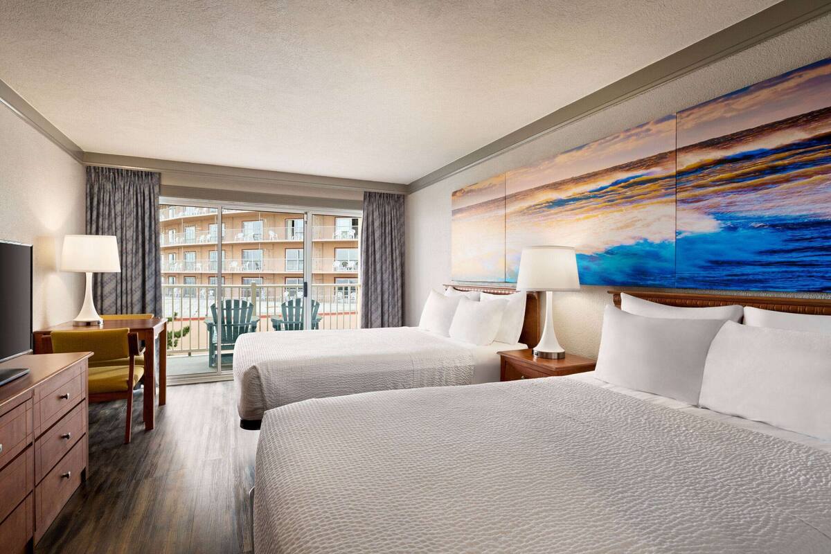 A hotel room with two beds, a desk, a chair, a TV, and a large window with a view of a building.