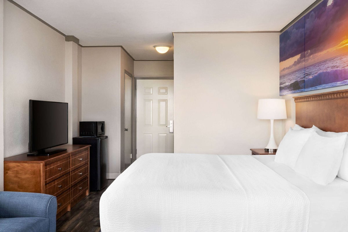 A hotel room with a bed, a TV, a dresser, and a painting of a sunset.