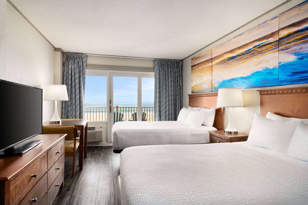 A hotel room with two beds, a TV, a desk, and a large window with a view of the ocean.