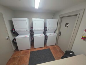 Un lavadero con tres lavadoras apiladas, un tapete negro en el suelo y una puerta con un letrero de cuarto de electricidad y calderas.