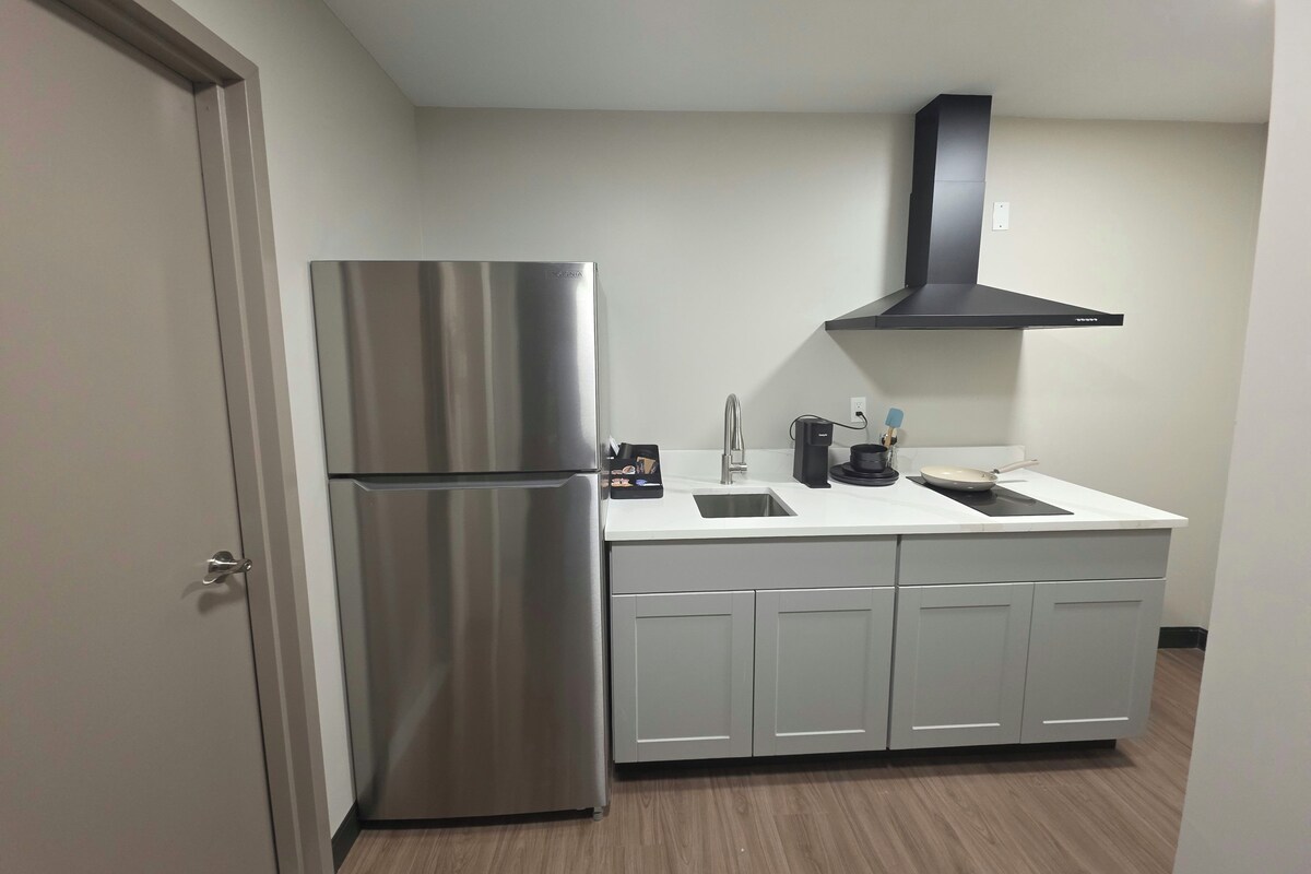 Una cocina compacta con refrigerador de acero inoxidable, campana extractora negra y encimera blanca.