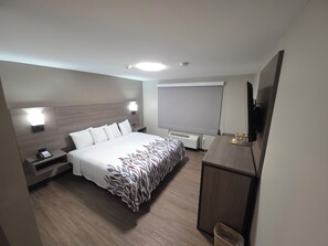 Habitación de hotel con cama, mesita de noche con teléfono, escritorio con computadora, televisor y ventana con persiana.