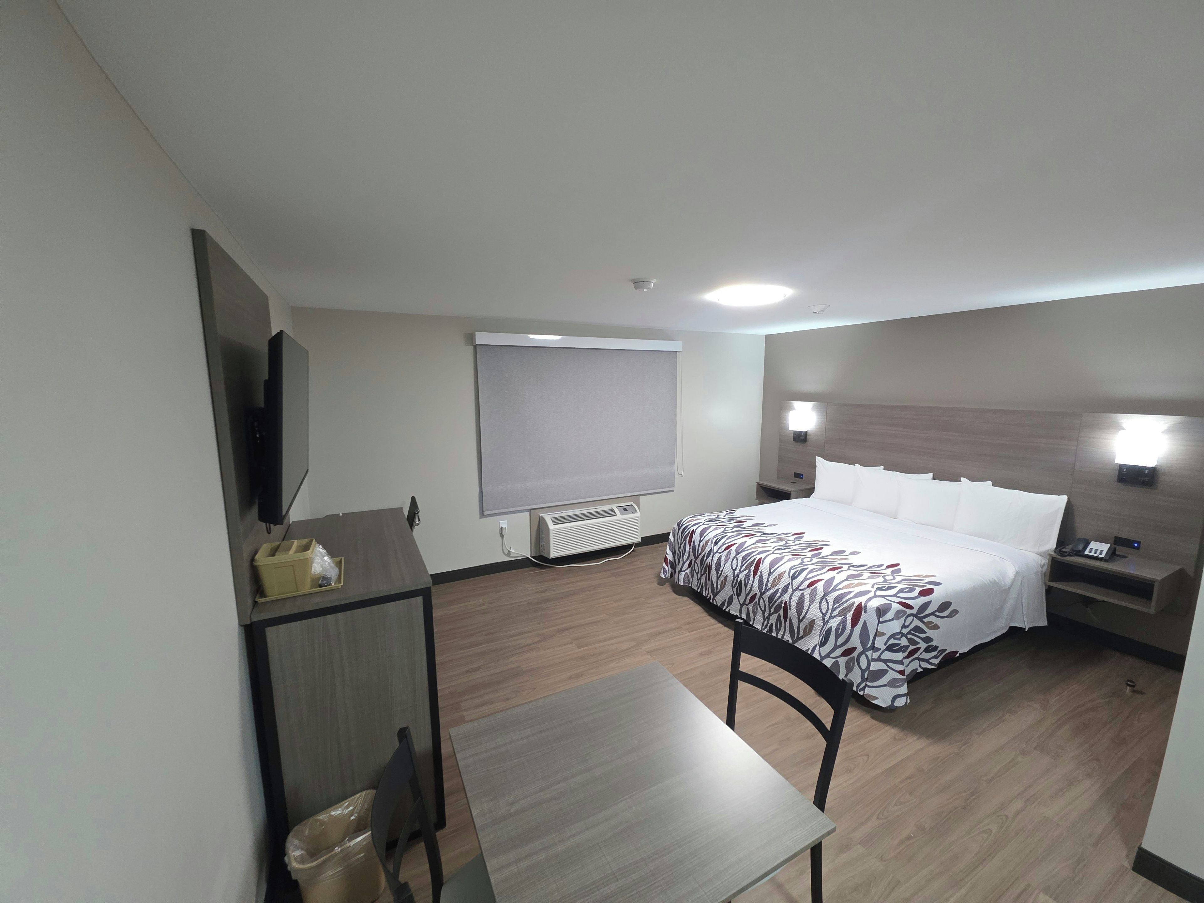 A hotel room with a bed, a desk, a chair, a TV, a window with blinds, and a bedside table with a lamp.