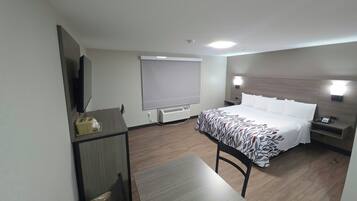 A hotel room with a bed, a desk, a chair, a TV, a window with blinds, and a bedside table with a lamp.