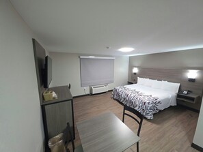 A hotel room with a bed, a desk, a chair, a TV, a window with blinds, and a bedside table with a lamp. - Red Roof Inn Sutton (Sutton)