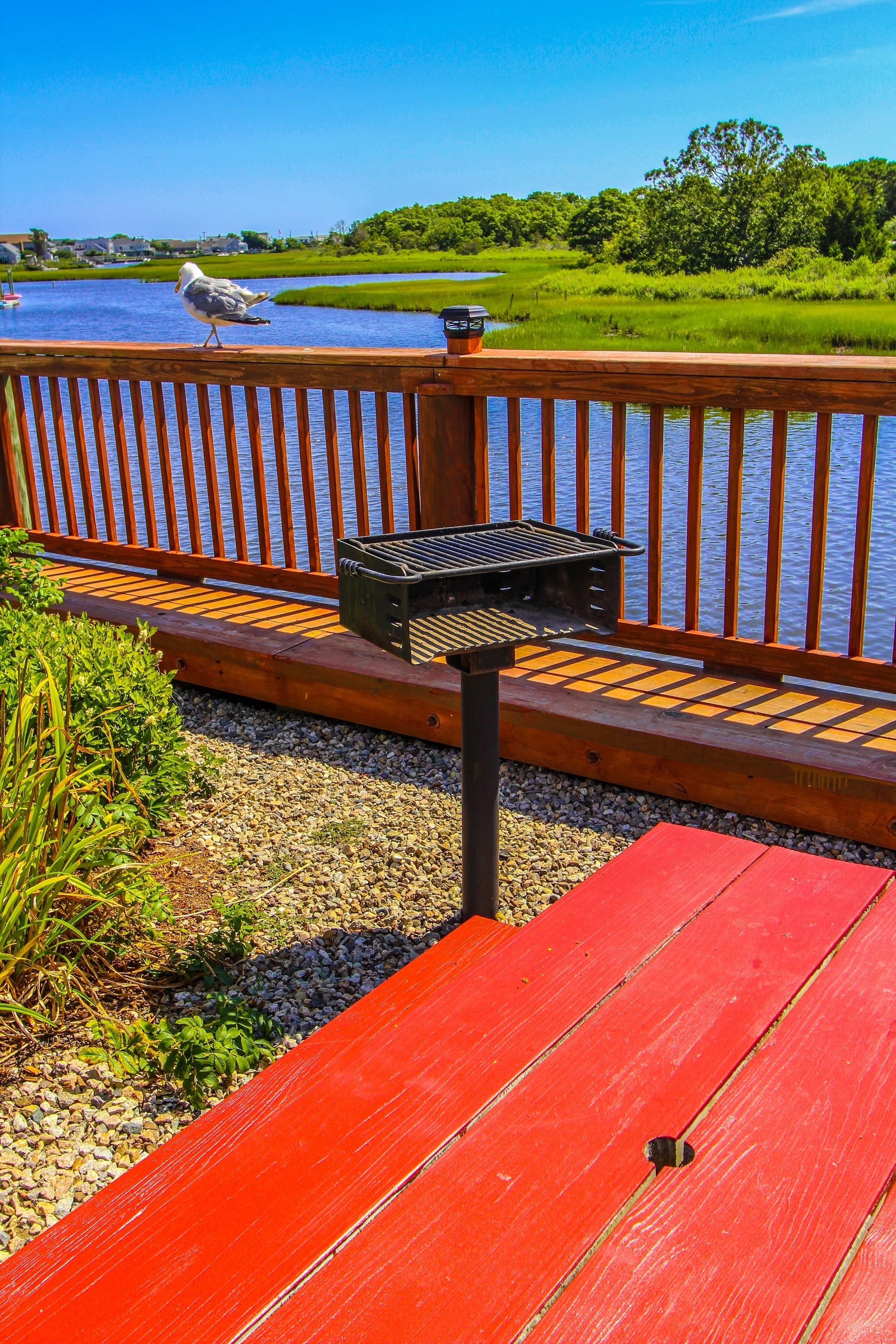 Une terrasse en bois avec un barbecue, un banc rouge et une vue sur un lac où l’on aperçoit un goéland.