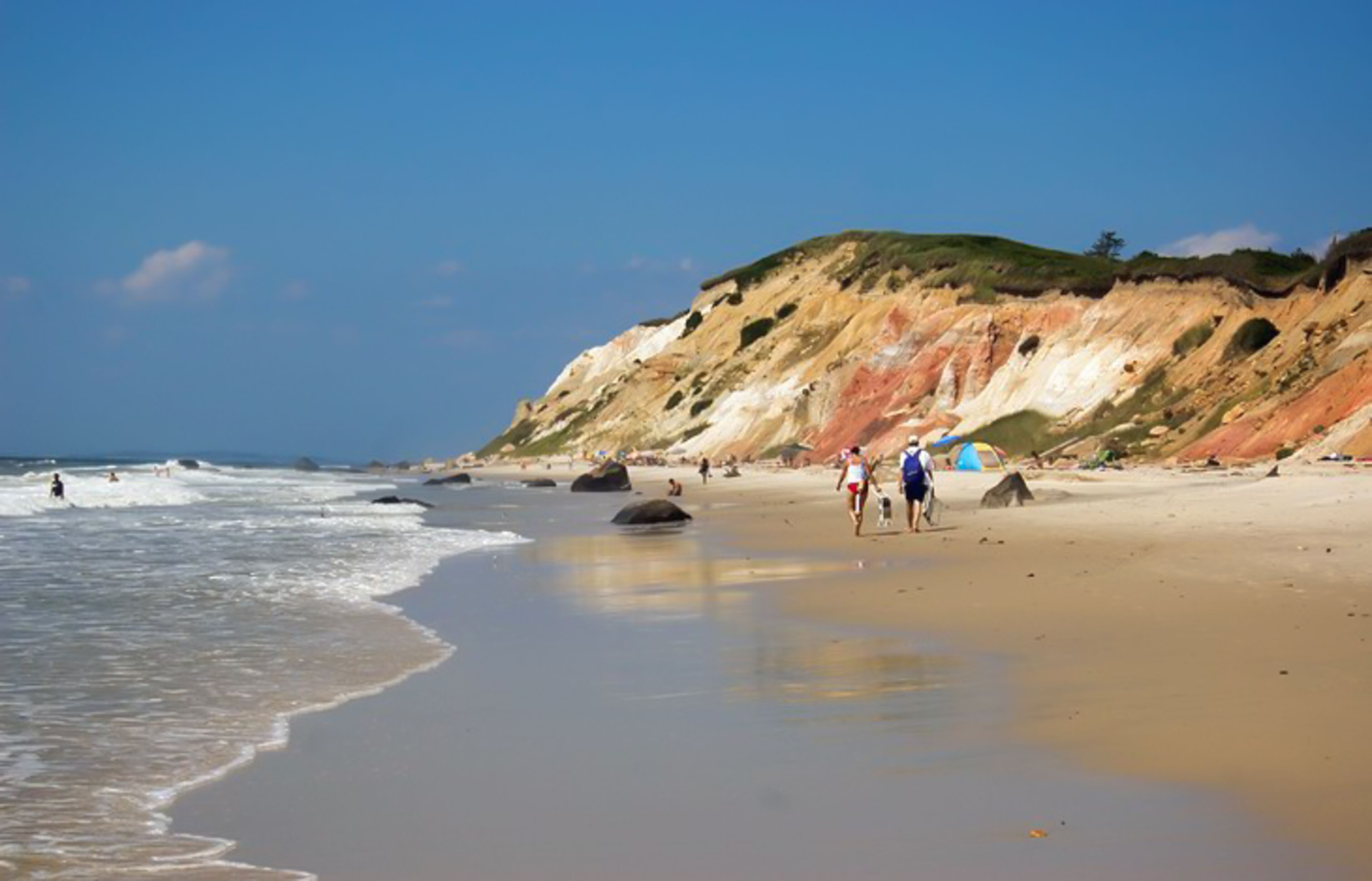 A beach with people walking along the shore, a sandy beach, and a rocky cliff in the background.