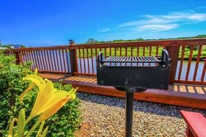 Eine Holzterrasse mit Grill aus Metall, die auf ein Gewässer blickt, umgeben von Grünpflanzen und einer gelben Blume.