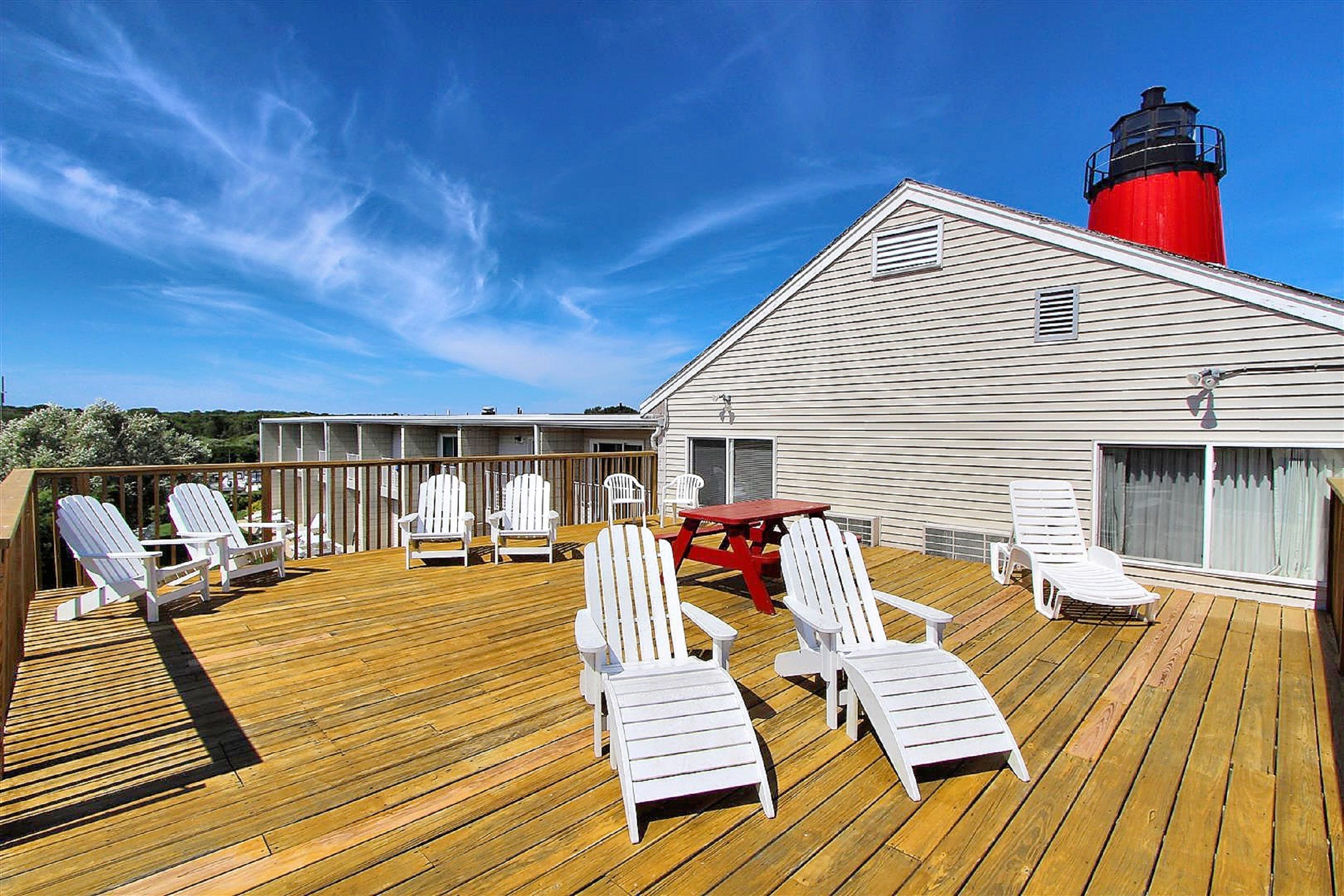 A deck with white Adirondack chairs, a red picnic table, and a red and black lighthouse in the background.