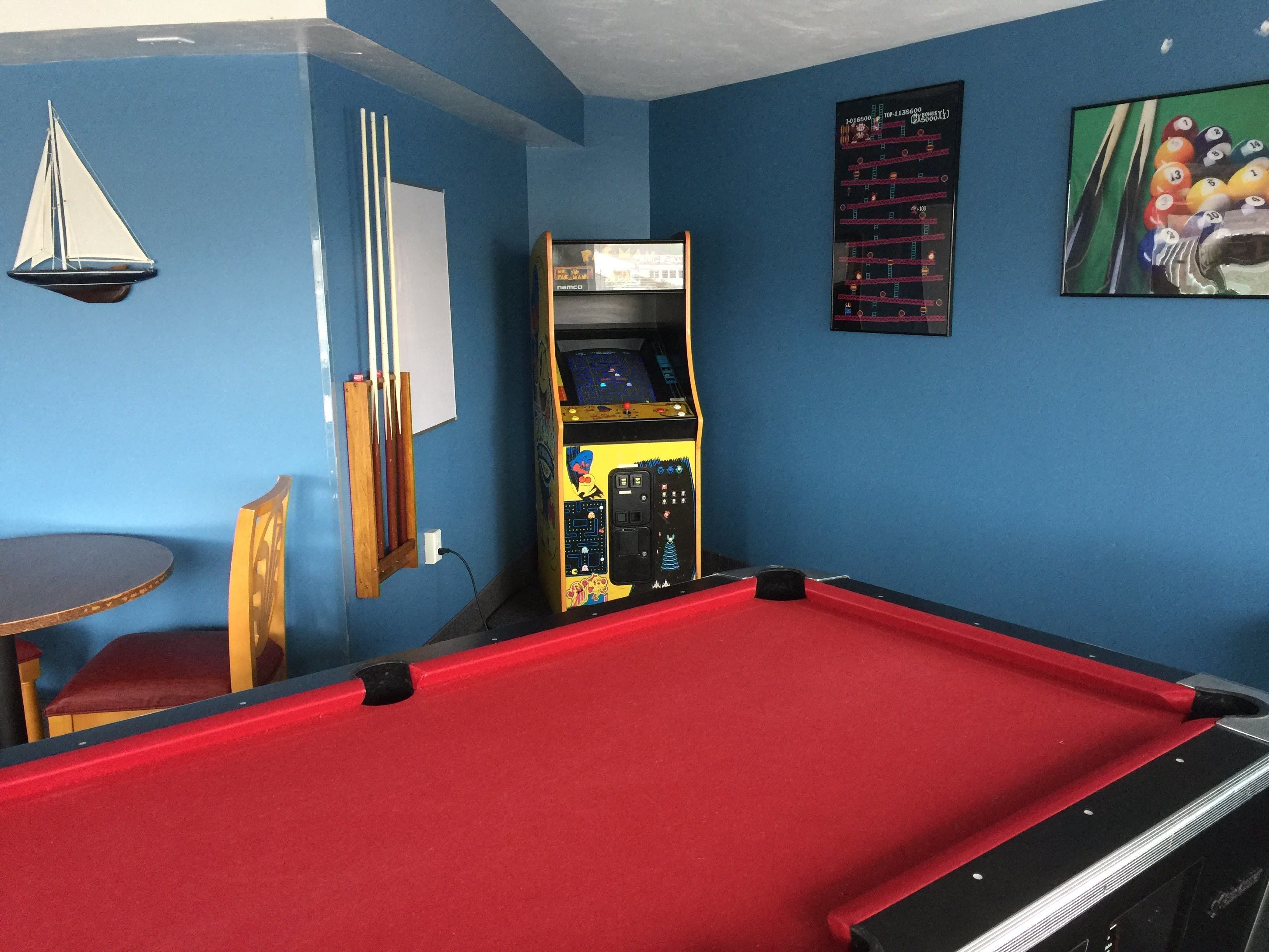 Une table de billard recouverte d’un feutre rouge, un porte-queue, une petite table avec un coussin et une borne d’arcade de billard.