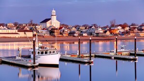 A waterfront marina with a white boat docked at a pier, a church with a steeple in the background, and residential buildings along the shore.