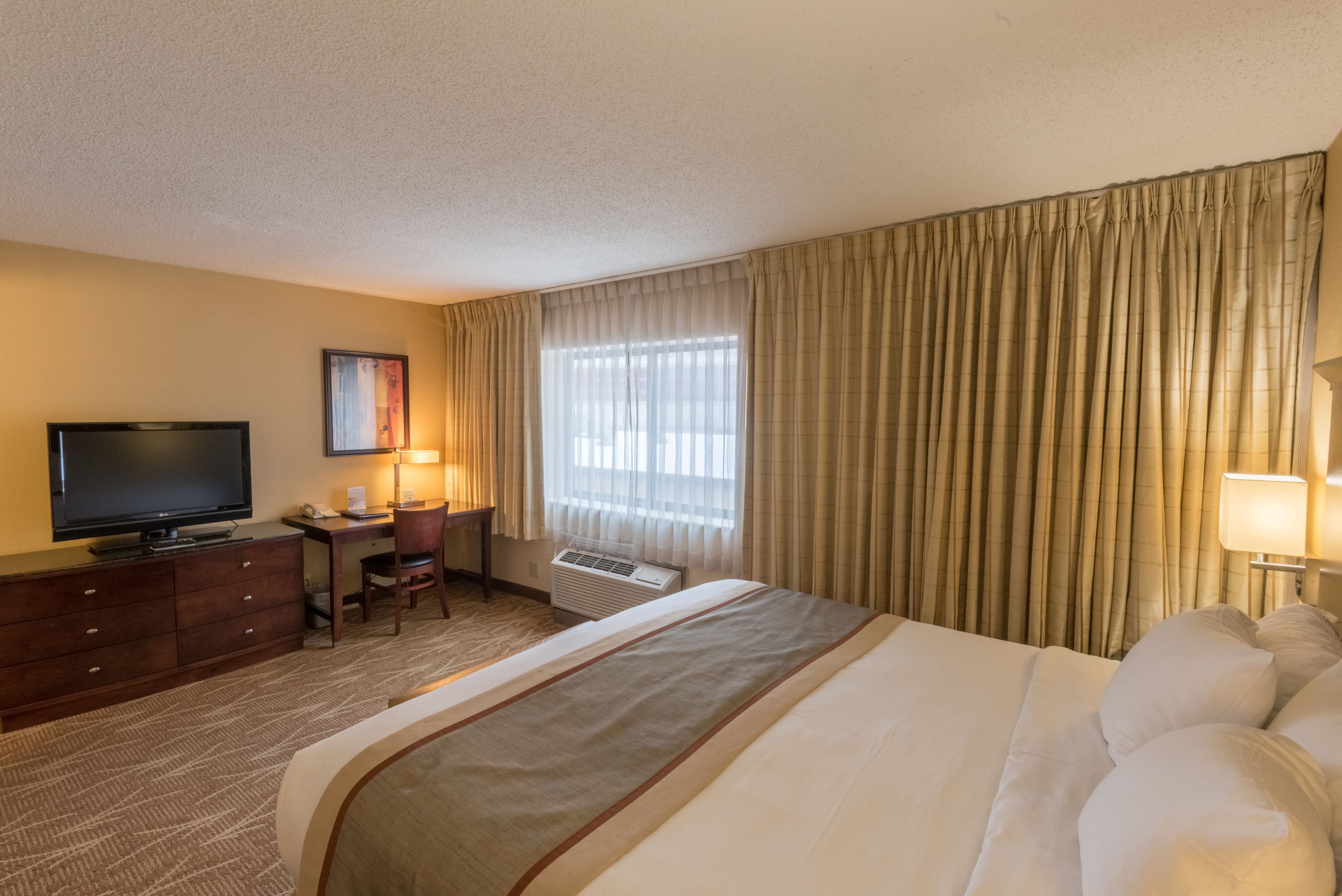 Ein Hotelzimmer mit Bett, Schreibtisch mit Computer, Fernseher und einem Fenster mit Vorhängen.
