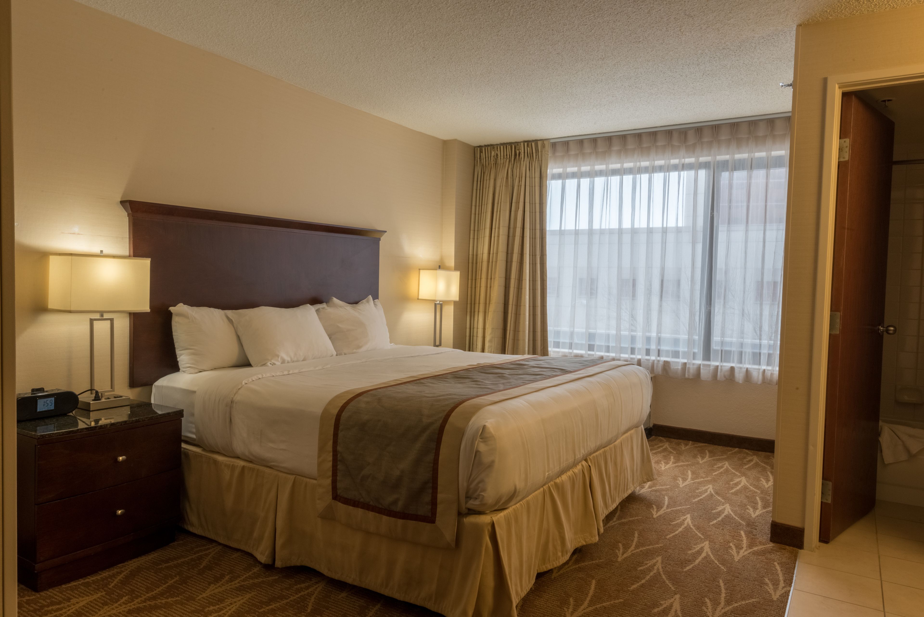 A hotel room with a large bed, two bedside lamps, a wooden headboard, a window with sheer curtains, and a carpeted floor.