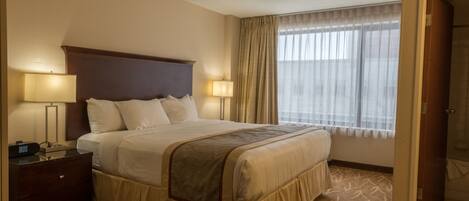 A hotel room with a large bed, two bedside lamps, a wooden headboard, a window with sheer curtains, and a carpeted floor.